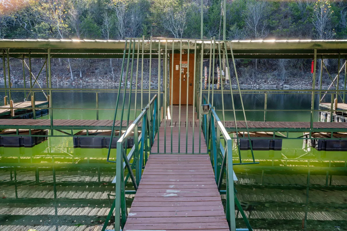 A peaceful lakeside dock with a wooden walkway and green railings leading to the water’s edge. The tranquil view highlights access to boating, fishing, and quiet moments by the lake, surrounded by natural greenery.