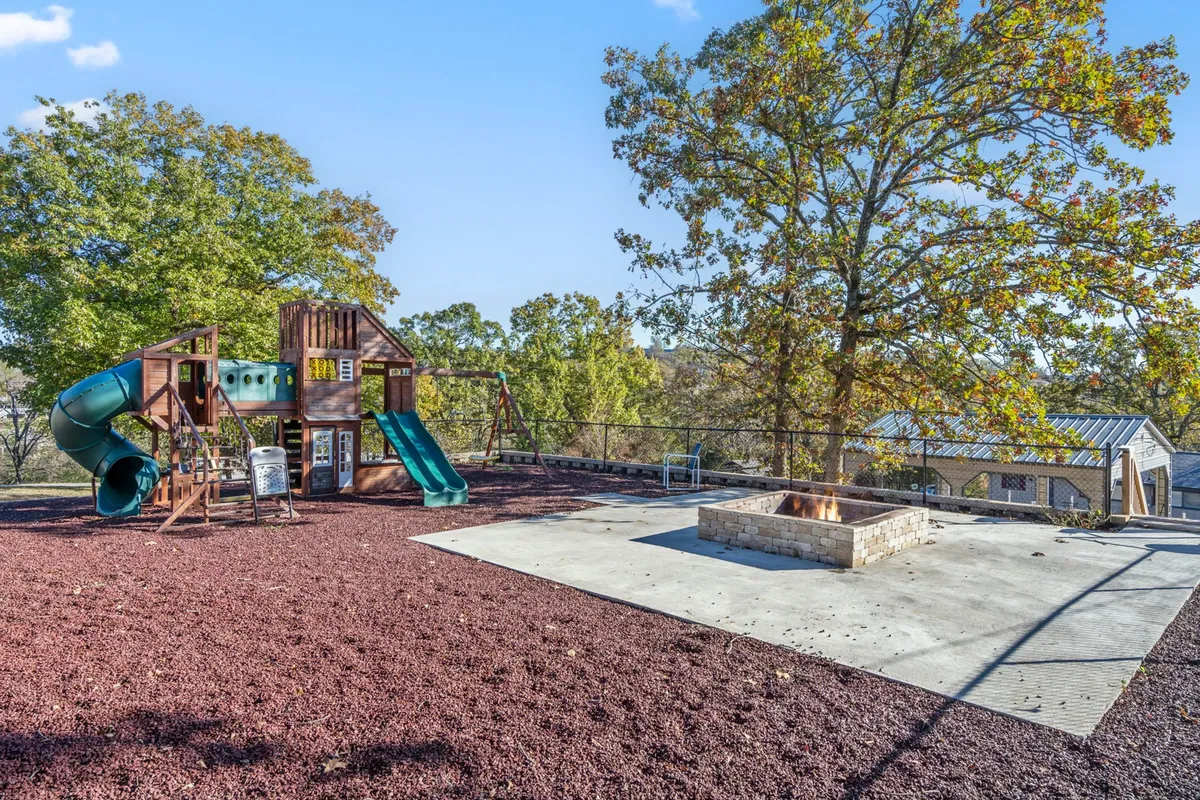 A lively recreation zone with colorful playground equipment and a modern fire pit, set among mature trees. The area is designed for both children’s play and cozy gatherings around the fire, with ample green space visible.