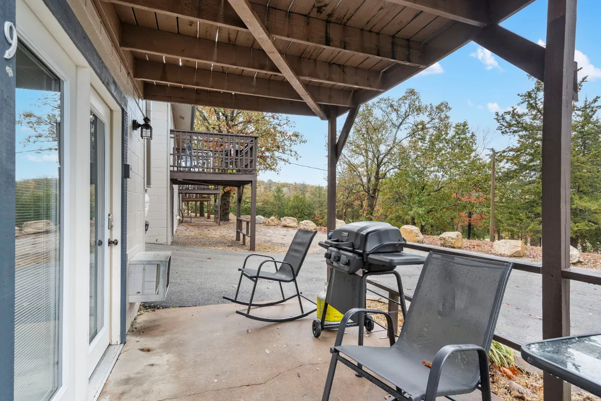 This image showcases a cozy exterior patio area featuring patio chairs, a grill, and sturdy wooden beams, all nestled among surrounding trees. The setup is inviting for both morning relaxation and evening gatherings, with abundant natural light and a tranquil backdrop.