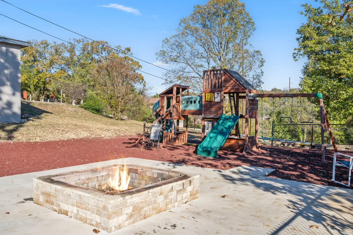 A family-friendly outdoor area featuring a playground structure and a fire pit, with ample greenery all around. The scene suggests both daytime play and evening gatherings in a cozy, safe environment.