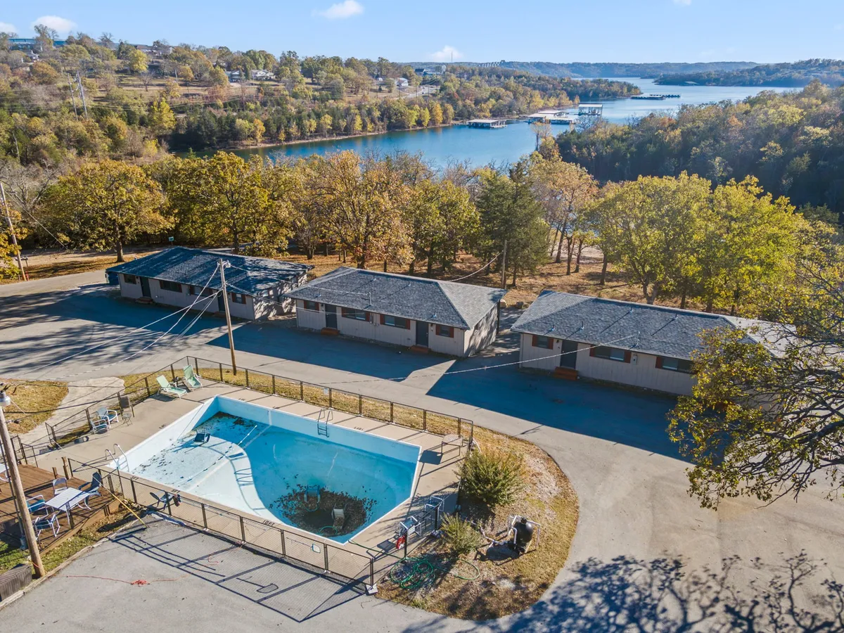A lively scene encompassing a swimming pool, shaded by trees, with a clear view of the lake and surrounding motel buildings. The space hints at on-site recreation like swimming, pool parties, and lakeside gatherings.