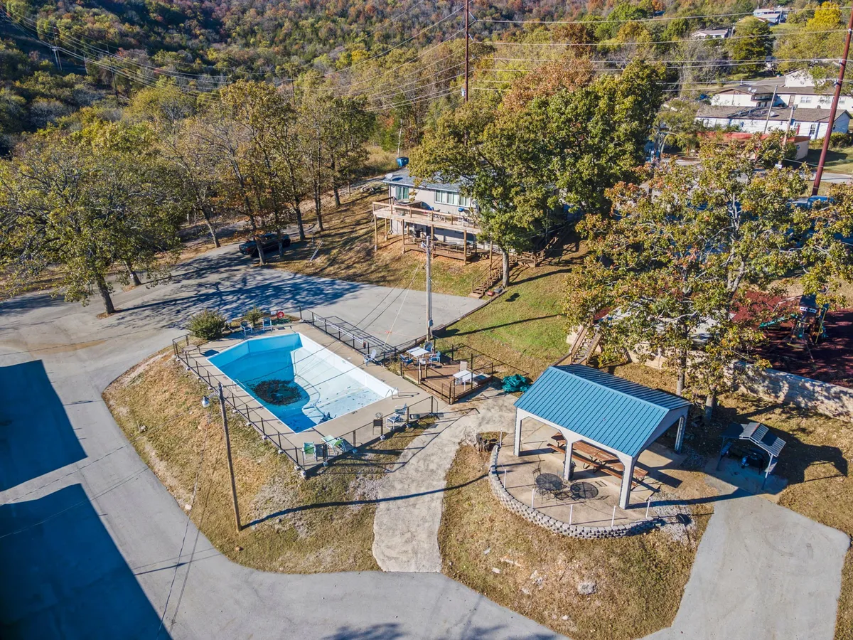 A vibrant outdoor pool scene featuring a sparkling swimming pool surrounded by lounge chairs and shaded gazebo seating, with leafy trees creating a lush backdrop. There's also an outdoor cooking area, evoking the feeling of summer relaxation and social gatherings.