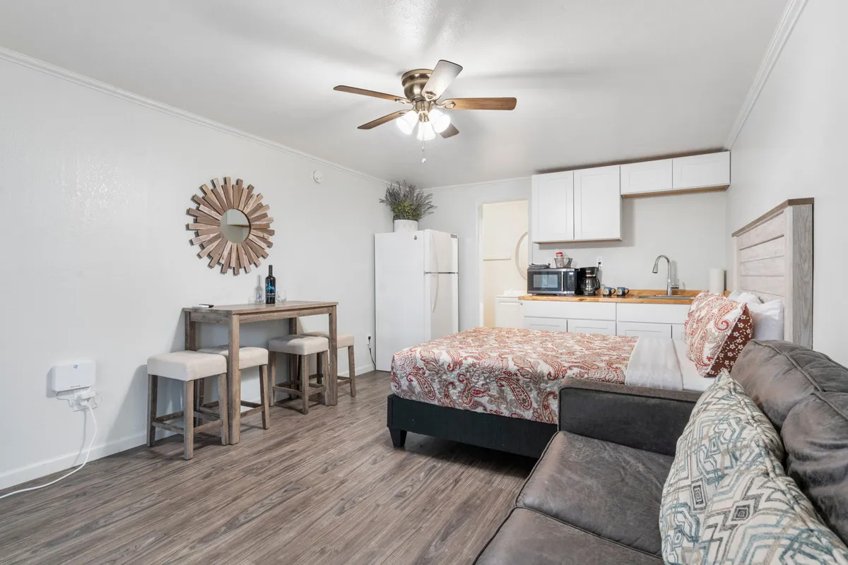 A cozy, homely studio interior featuring a bed with patterned bedding, a small dining table with stools, a comfortable sofa with decorative pillows, and a wall mirror. Sunlight pours in through large windows, enhancing the sense of space and warmth.