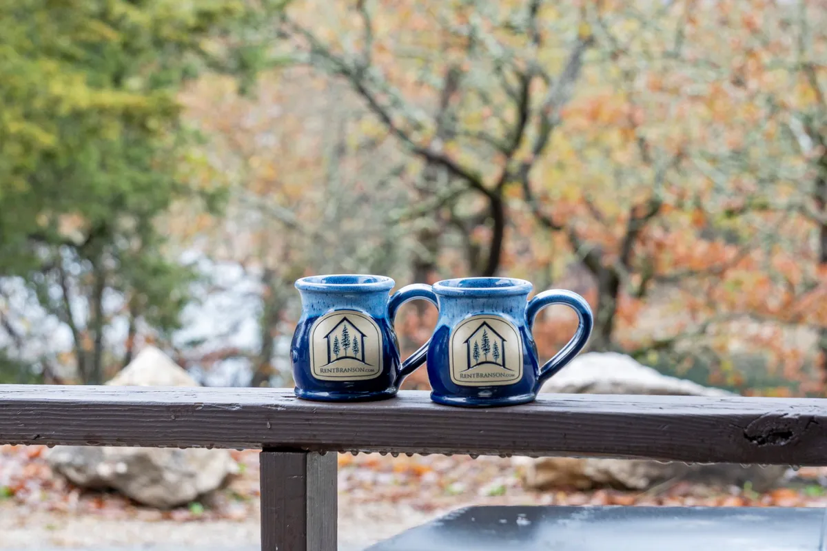Here, two ceramic mugs rest on a wooden patio railing overlooking a lush natural background. The peaceful setting emphasizes intimacy and connection with nature, hinting at the enjoyment of fresh air and lake sounds mentioned in the content.