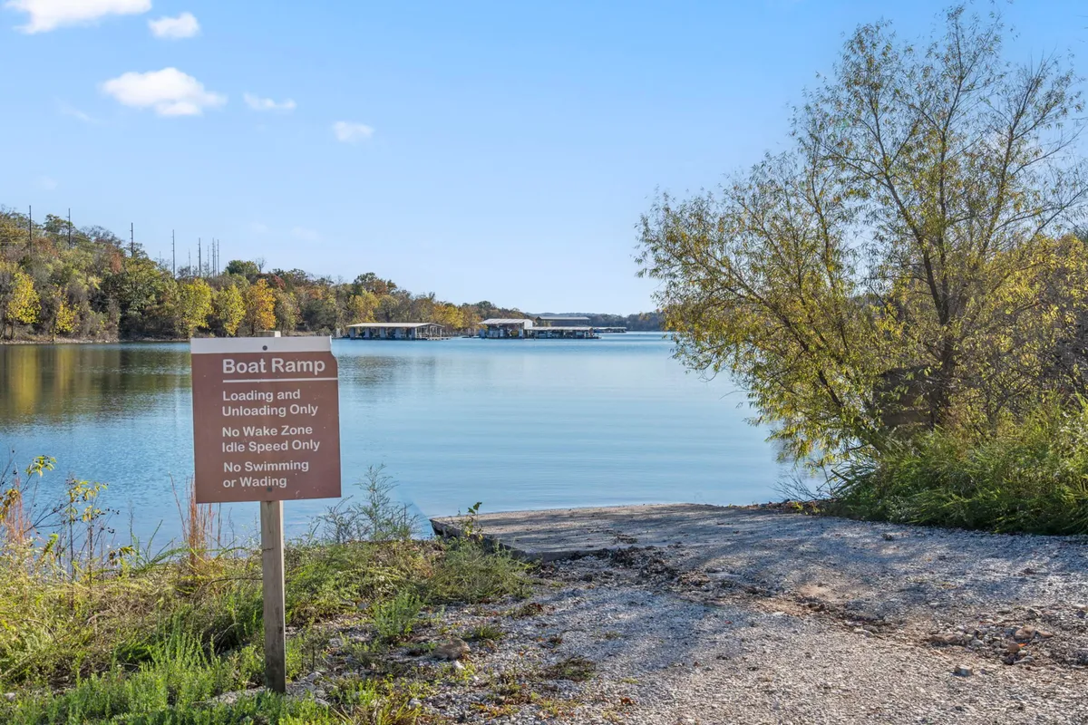 A tranquil lakeside scene featuring calm water, a boat ramp sign, surrounding trees, and distant boat docks. The sun appears low, hinting at sunrise or sunset, with the lakeshore inviting morning strolls or peaceful evening reflection.